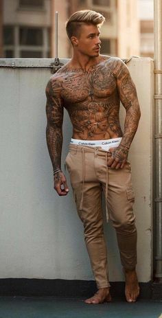 a tattooed man leaning against a wall with his hands on his hips wearing khaki pants