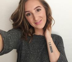 a woman with a tattoo on her arm pointing at the camera while wearing a gray shirt