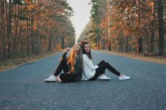 two young women sitting on the side of a road in the middle of an autumn forest