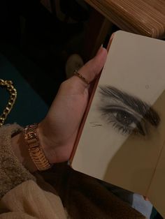 a woman holding an open book with her eye drawn on the page and wearing gold bracelets