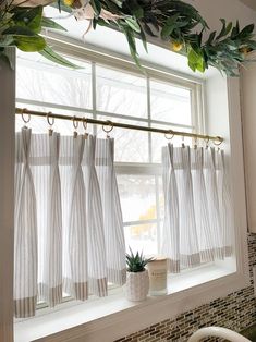 a kitchen window with white curtains and green plants hanging from it's rodulage