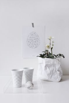 two white vases sitting on top of a table next to a plant in a bag