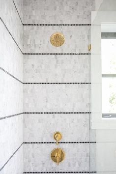 a white tiled shower with gold faucet and hand held shower head next to window