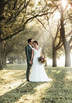 a bride and groom standing in the grass under trees with sunbeams behind them