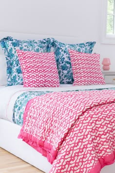 a white bed topped with pink and blue comforter next to a window covered in pillows