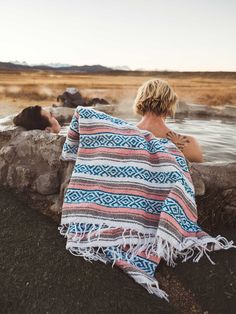 a woman wrapped in a blanket sitting next to a body of water