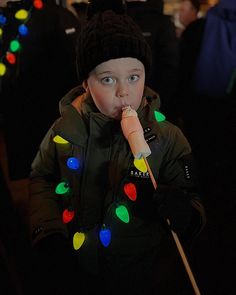 a young boy wearing a hat and jacket with christmas lights around his neck, eating an ice cream cone