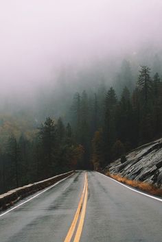 an empty road in the mountains with trees on both sides and foggy skies above