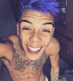 a young man with blue hair and tattoos on his chest smiling at the camera while standing in a kitchen