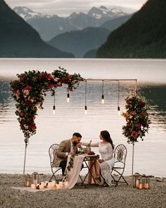 a man and woman sitting at a table on the beach with candles in front of them