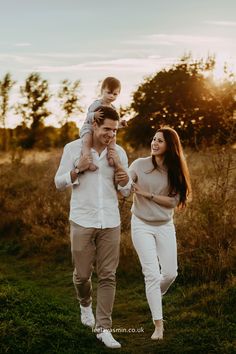 a man and woman walking through a field with a baby on their shoulders, while the sun is setting behind them