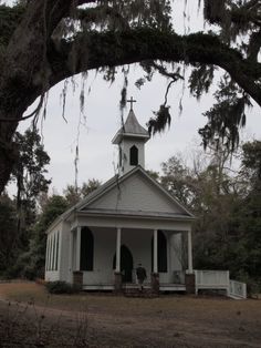 an old white church with a steeple surrounded by trees and mossy branches on a cloudy day