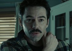 a man with a moustache stares into the camera while standing in front of a window