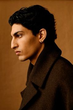 a man with black hair wearing a coat and looking off to the side in front of a brown background