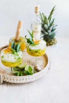 two glasses filled with drinks sitting on top of a wooden tray next to pineapples