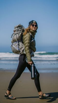 a woman walking on the beach with a backpack and water bottle in her hand as she walks towards the ocean