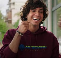 a young man with curly hair wearing a maroon hoodie smiles and holds his fist up