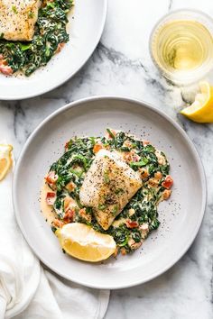 two white plates topped with fish and spinach next to lemon wedges on a marble table