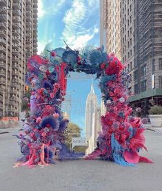 an arch made out of flowers and feathers in the middle of a street with tall buildings behind it