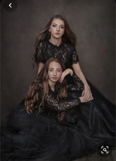 two young women in black dresses posing for a photo together, one is sitting on the floor