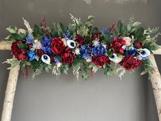 an arrangement of red, white and blue flowers on a wooden arch in front of a gray wall