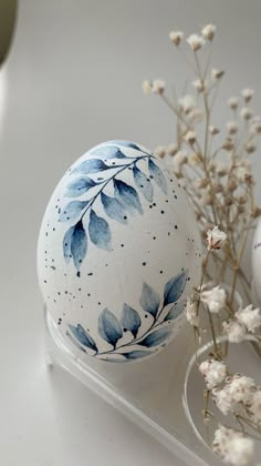 an egg with blue leaves painted on it next to some white flowers and a glass vase