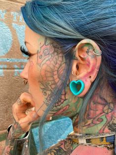 a woman with blue hair has tattoos on her face and behind her ear is a heart