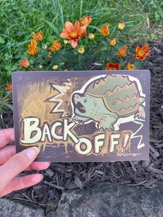 a hand holding up a sticker with an image of a dinosaur and the words back off