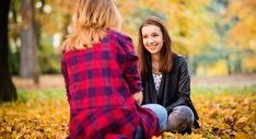 two young women sitting on the ground talking to each other in an autumn park full of leaves