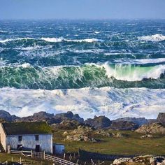 an old house sitting on the shore with waves crashing in front of it and a large body of water behind it