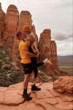 a man and woman standing on top of a rock formation with mountains in the background