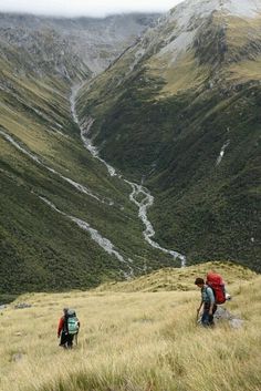 two people with backpacks are hiking up a hill side in the mountains near a river