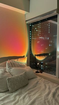 a bed with white sheets and pillows in front of a window that has city lights on it