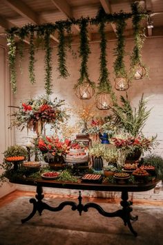 a table topped with lots of food and greenery next to a wall filled with hanging lights