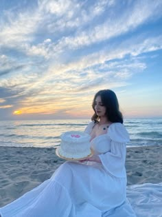 a woman sitting on the beach with a cake in front of her and sunset behind her