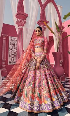 a woman standing in front of a pink wall wearing a colorful lehenga set