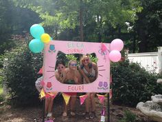 three girls are posing in front of a pink frame with balloons and cat ears on it