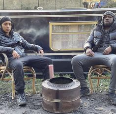 two people sitting on chairs next to an open fire pit in front of a train