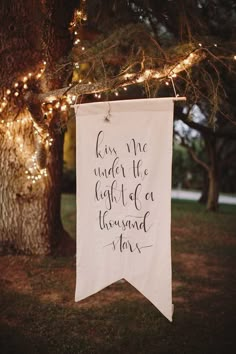 a white banner hanging from a tree with string lights behind it that says, kiss me under the light of our thousand stars