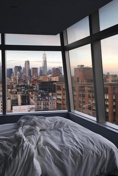 an unmade bed in front of large windows overlooking cityscape and skyscrapers