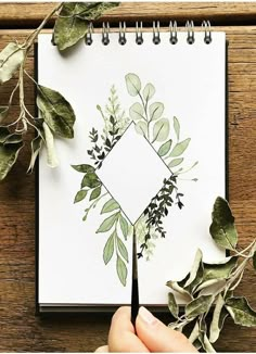 a hand holding a pencil over a notebook with leaves and plants around it on top of a wooden table