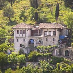 an old house on the side of a hill with lots of trees and bushes around it