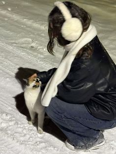 a woman kneeling in the snow petting a small white and brown cat with headphones on