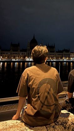 two people sitting on the edge of a pier at night looking out over water with buildings in the background