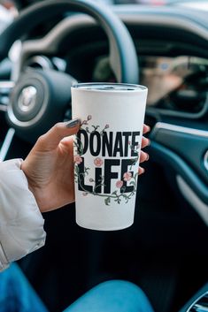 a person holding up a coffee cup with the words donation life printed on it in front of a car steering wheel