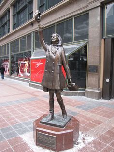 a statue of a woman holding an umbrella in the middle of a city street with people walking by