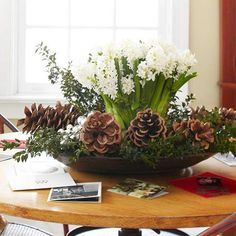 a wooden table topped with a bowl filled with flowers and pineconis next to a window