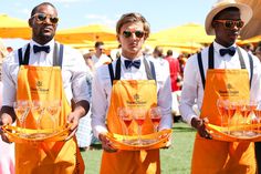 three men in orange aprons holding trays with wine glasses on them and yellow umbrellas behind them