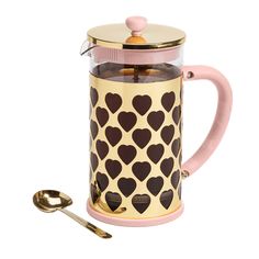 a pink and gold coffee pot with hearts on it next to two spoons,