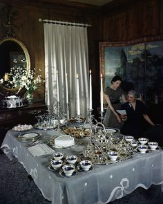 a man and woman standing next to a table covered in silver dishes, cups and saucers